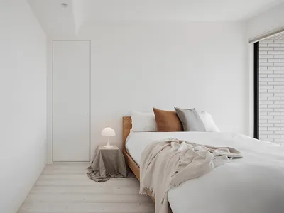 A minimalist bedroom with a white bed, wooden frame, and a bedside lamp on a beige fabric-covered table.