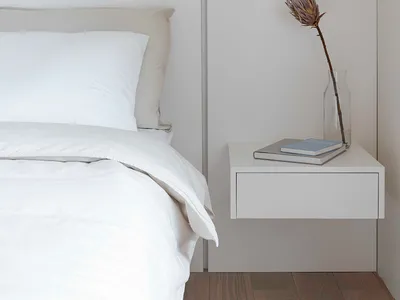 A serene, minimalist bedroom featuring a white bed with neatly arranged white bedding and a simple, white nightstand with a v