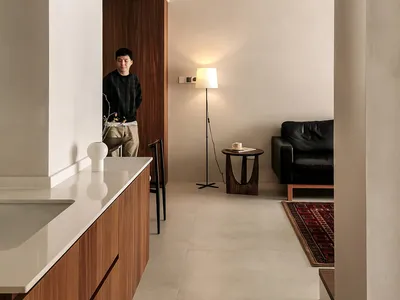 極簡客廳，鋪設混凝土地板，搭配黑色皮革沙發，以及小型木質邊桌，桌上擺放燈具與杯子