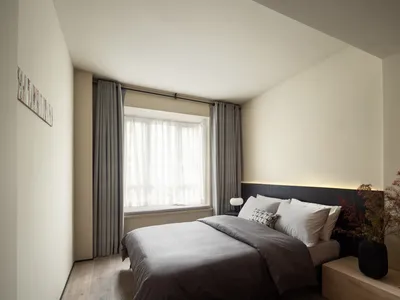 A minimalist bedroom with neutral-colored walls, curtains, and bedding, featuring a large window and a cozy bed setting.