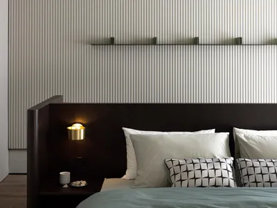 A modern minimalist bedroom with a wooden headboard, fabric bedding, and metallic accents on the nightstand and shelves.