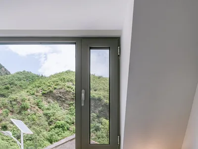 A minimalist bedroom featuring a bed with white sheets and a wooden headboard, overlooking a green hillside through a large w