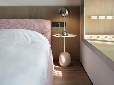 A minimalist bedroom with a pink headboard bed, white bedding, and a wooden nightstand with a round table lamp.