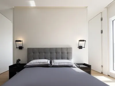 Modern minimalist bedroom with a grey upholstered headboard, white bedding, and black sconces on either side of the bed.
