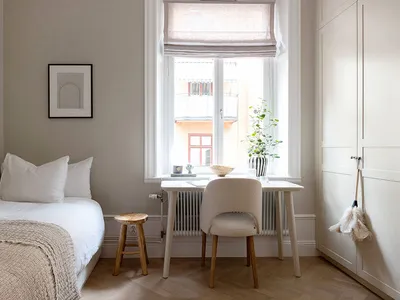 Bedroom filled with light, neutral tones, featuring a bed, a desk, a stool, and a window with a plant.