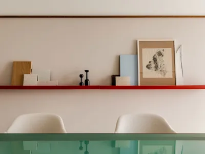 A minimalist home office with a red shelf displaying various objects, including a framed abstract piece, small sculptures, an