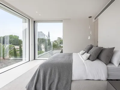 A modern, brightly lit bedroom with a bed dressed in light fabrics and glass windows.