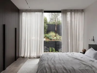 A minimalist bedroom featuring white curtains, a wooden floor, and a white bed with a view of greenery outside.