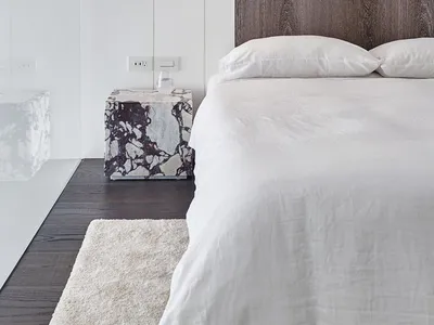 Modern minimalist bedroom featuring a wooden headboard against a white wall, illuminated by a wall-mounted sconce, with a whi