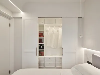 A minimalist bedroom with white walls, a bed, and a built-in closet with white doors and white hanging garments.