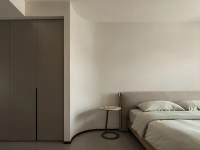 A minimalist bedroom with neutral-colored furniture and walls, featuring a bed with a low-profile headboard and a simple side