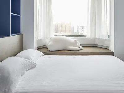 A minimalist bedroom featuring a white bed with cushions and a white stuffed animal, positioned in front of a large window wi