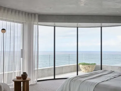 A modern minimalist bedroom featuring a bed in the foreground, with a large window overlooking the ocean, and light-colored c