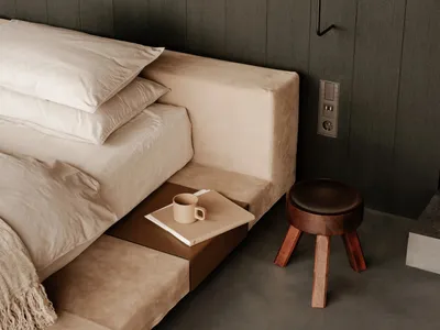 A minimalist bedroom featuring neutral-toned furniture, a modern side table with a coffee mug and book, and a sleek, dark-col