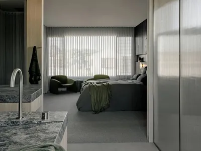 Bedroom with concrete flooring, a large window with vertical blinds, a green armchair, and a bed covered in a green blanket.