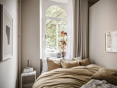A cozy minimalist bedroom featuring a bed dressed in beige textiles, a small side table, and a window with white curtains ove