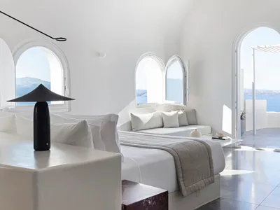 Modern, minimalist bedroom with white walls and concrete floor, featuring a cozy bed and seating area with arched windows.