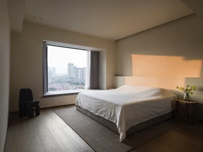 A minimalist bedroom with a large window providing a view of the cityscape, featuring a bed with a white sheet and a wooden s