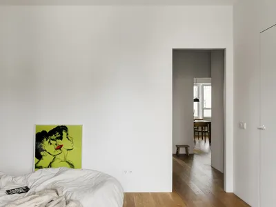 An image of an empty minimalist bedroom with white walls, wood flooring, and a green and yellow framed artwork.