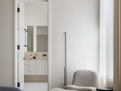 Modern bedroom featuring a neutral color palette with a wooden bench, a plush grey chair, and a minimalist bathroom seen thro
