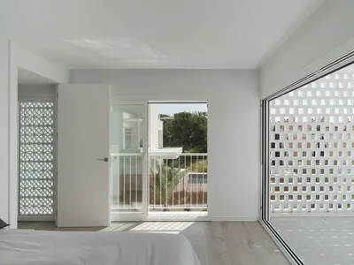 A minimalist bedroom with white walls, wood-laminate flooring, and a large window overlooking a balcony with a metal railing.