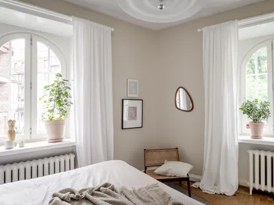 A minimalist bedroom with white curtains and walls, featuring a bed with a soft blanket, a small table with a lamp, and potte