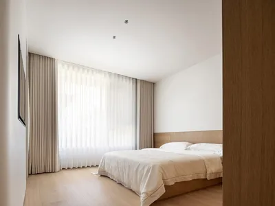 Minimalist bedroom with a bed, wooden furniture, light textiles, plastic blinds, white walls, and a wooden floor.