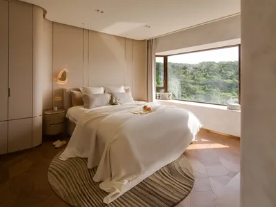 A modern bedroom with white cushions on the bed, a large window with a view of greenery, and a minimalist design.