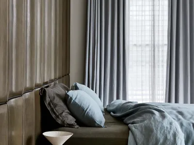 A modern minimalist bedroom with fabric headboard, light grey curtains, and light blue bedding.