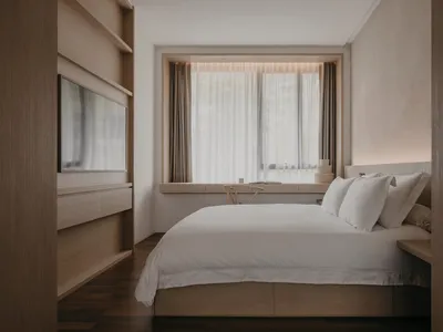 Modern minimalist bedroom with a large window, white bedding, and wood furniture.