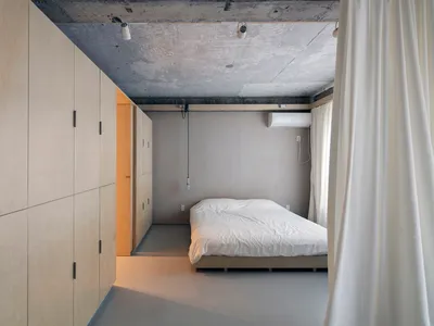 A minimalist bedroom with a concrete floor and walls, featuring a plain white bed with a simple wooden frame.