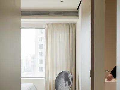 A modern minimalist bedroom with white walls, white curtains, a white bed, and a circular speaker with a moon pattern on a wo