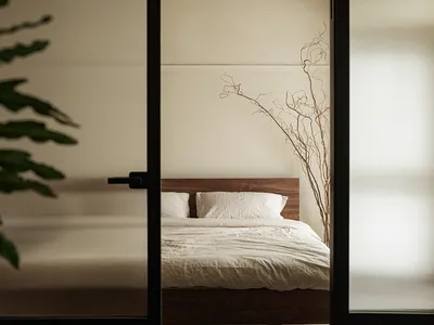 Minimalist bedroom with a wooden bed frame and white bedding, viewed through a glass door with green foliage outside.