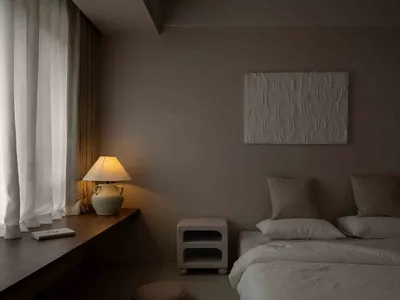 Minimalist bedroom with neutral tones, featuring a bed, nightstand, lamp, and artwork