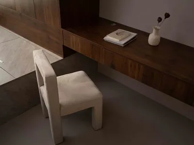 A minimalist home office desk made of wood with a stack of books and a vase on top, and a beige chair.
