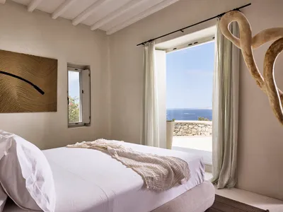 A minimalist bedroom with neutral tones, featuring a white bed, light-colored curtains, and a view of the ocean through the w