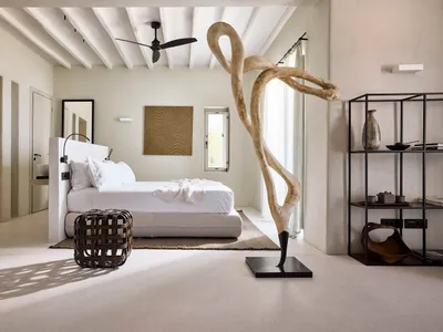 A minimalist bedroom featuring a bed with neutral bedding, a large abstract sculpture, and a modern bookshelf.