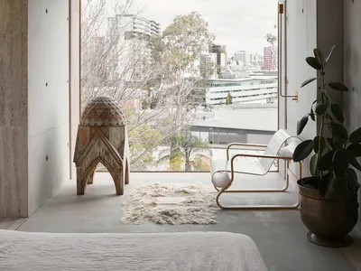 A minimalist bedroom with concrete walls, a white bed, a white chair, and a plant in a large window with a city view.