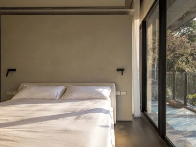 A modern minimalist bedroom with a wooden floor, white linens, and a large window overlooking greenery.