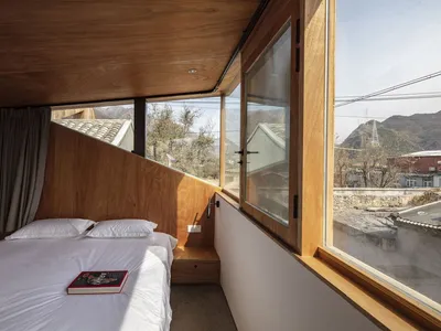 A modern bedroom with a bed, pillows, and a book on a wooden floor, large windows, and a view of the mountains.