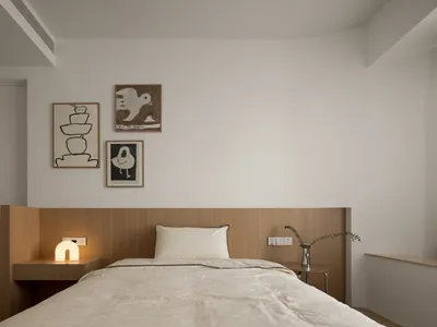 A minimalist bedroom with a wooden bed frame, white bedding, and three framed artworks on the wall.