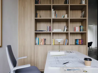 A modern home office with wooden shelves containing books and a white desk with blueprints and a coffee cup on it.