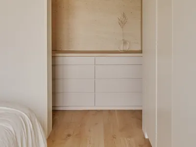 A minimalist bedroom storage unit featuring neutral-toned drawers and a vase with a dried plant against a natural wood floor