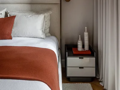 A modern minimalist bedroom with a grey upholstered headboard, orange throw blanket, white bedding, and fabric curtains in th