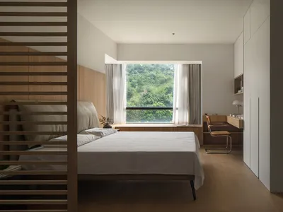 A minimalist bedroom featuring a bed with white sheets and a wooden headboard and floor, with a large window showcasing a lus
