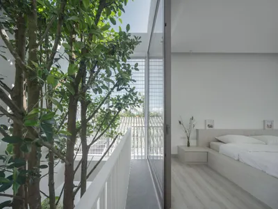 A minimalist bedroom with a white bed and wooden floor, featuring green trees outside the large windows and a clean, modern d