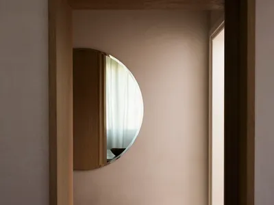 A minimalist bedroom featuring a circular mirror on the wall, a wooden bench, and sheer curtains, creating a serene and moder