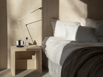 A minimalist bedroom featuring a bed with white bedding, a wooden nightstand with a lamp, and wooden walls.