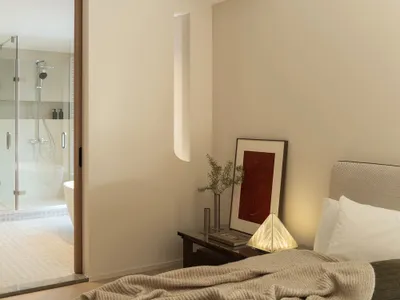 A minimalist bedroom featuring neutral-toned walls, a bed with a modern, textured headboard, and a lighted table lamp.