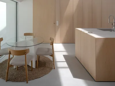 A modern minimalist residential kitchen with wood cabinetry, a glass dining table, and a large window allowing natural light.